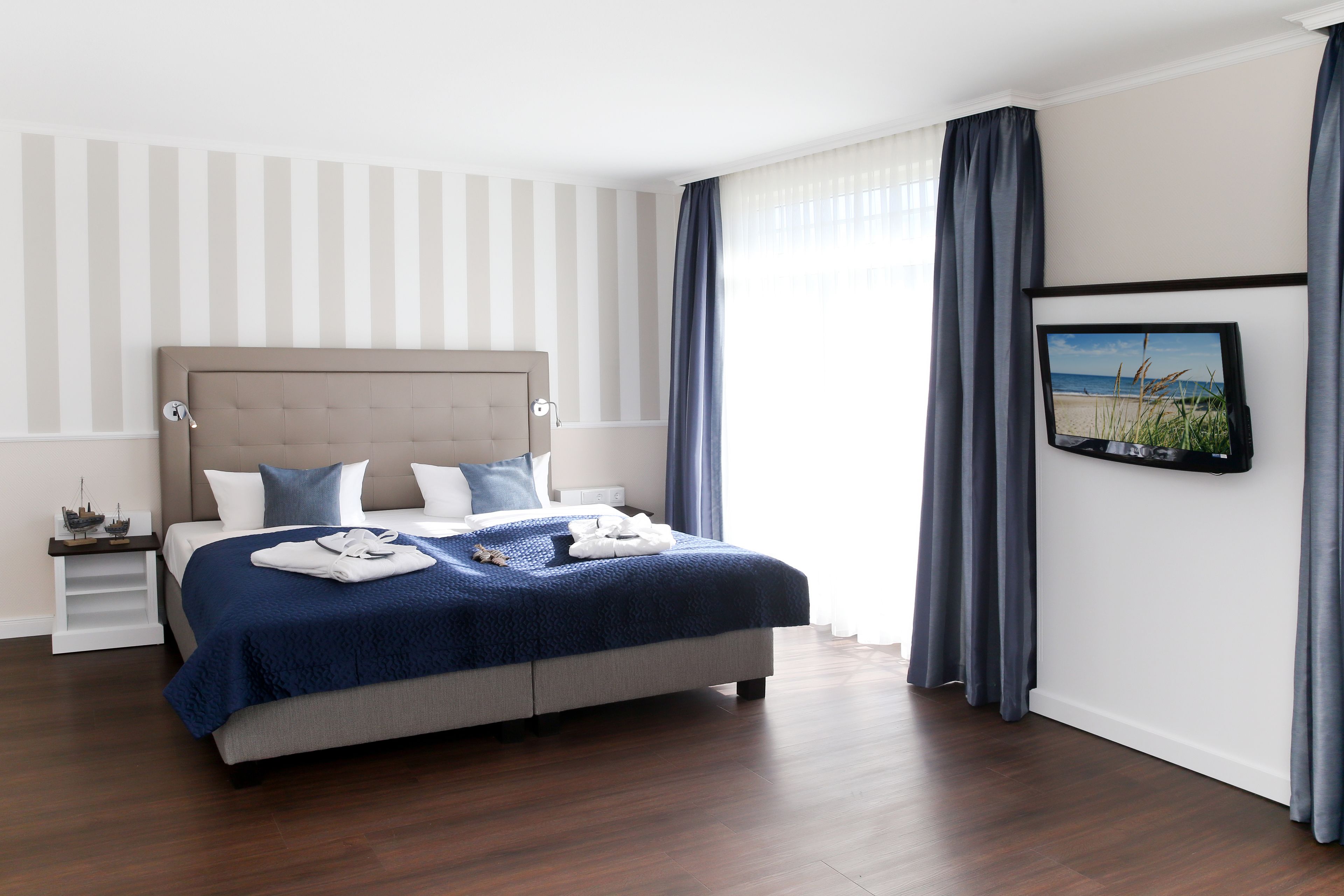 Een netjes ingerichte slaapkamer met een groot bed, een nachtkastje, een aan de muur bevestigde televisie en een raam met gordijnen.