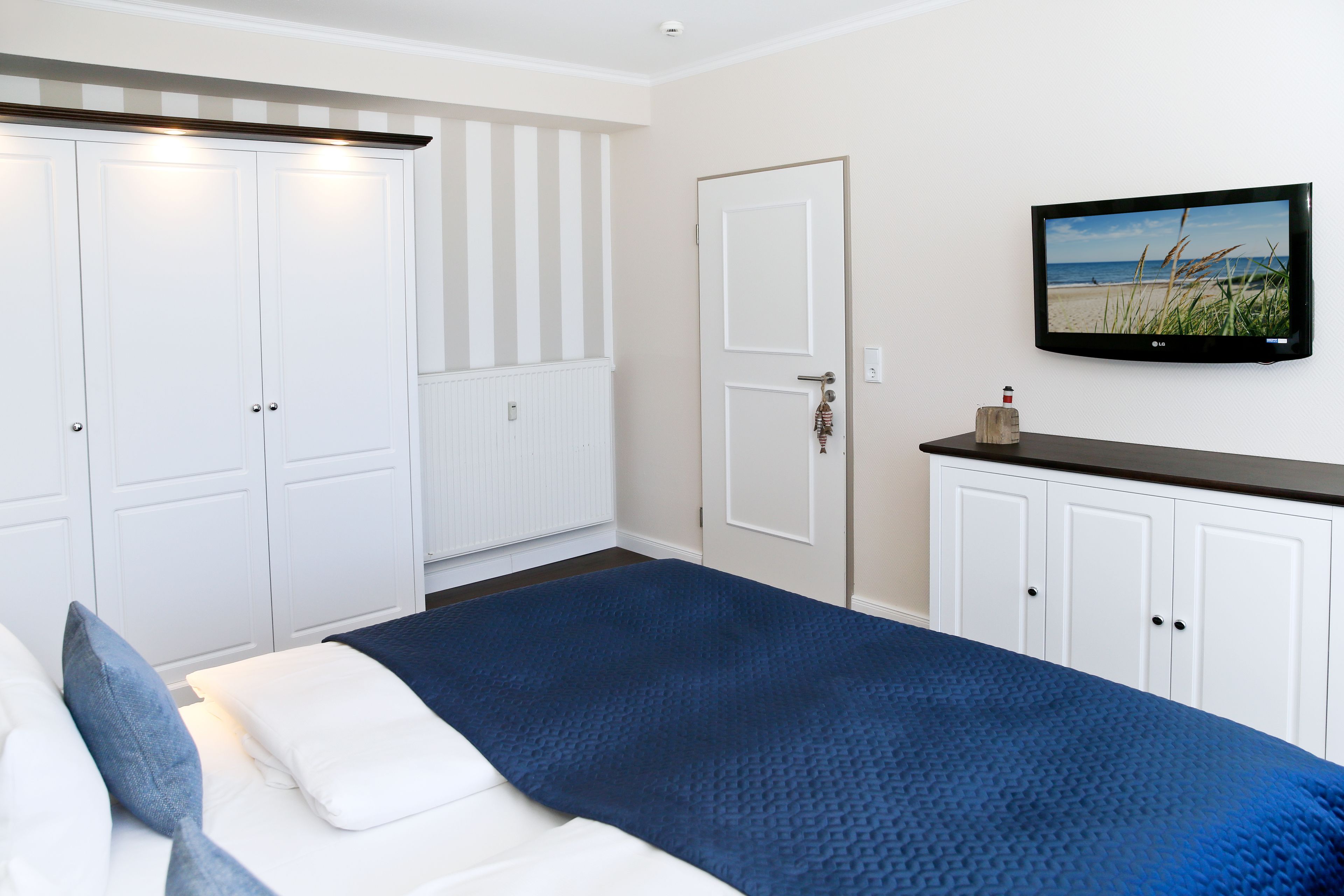 A bedroom with a bed, white wardrobes, a television on the wall, and a beach scene on the screen.