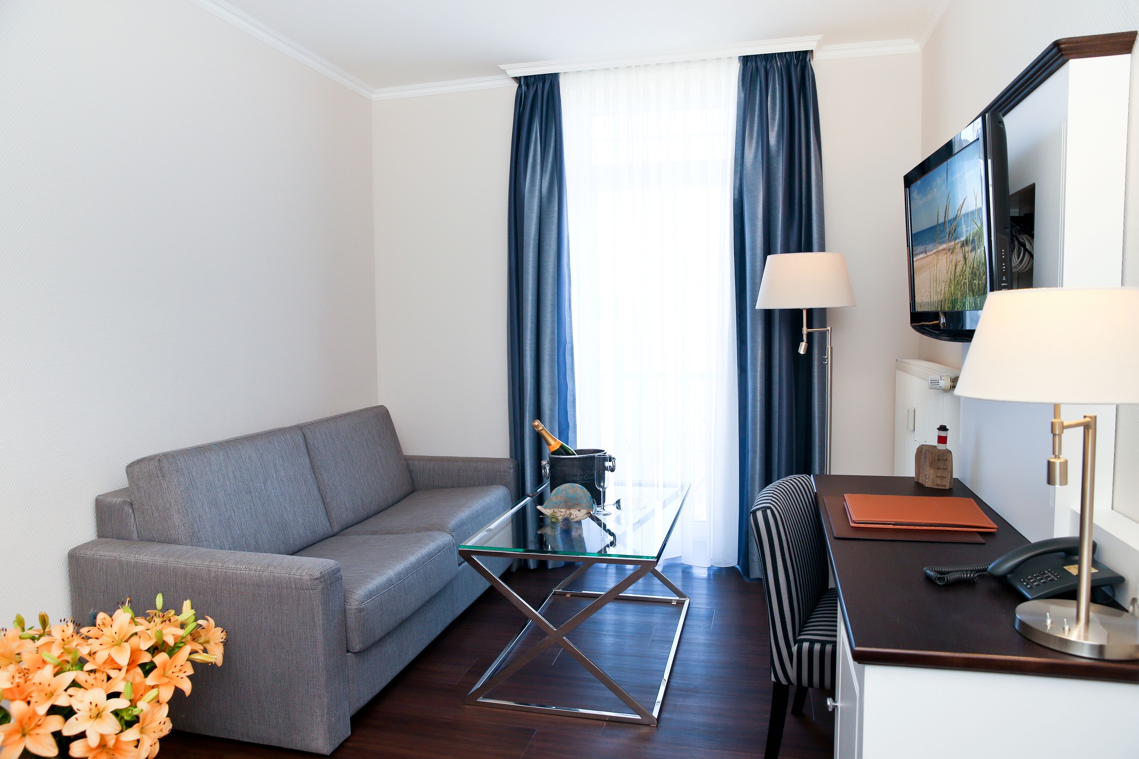 A modern hotel room with a grey sofa, a glass coffee table, a wooden desk with a lamp, a television, and a view of the outdoors through large windows with blue curtains.