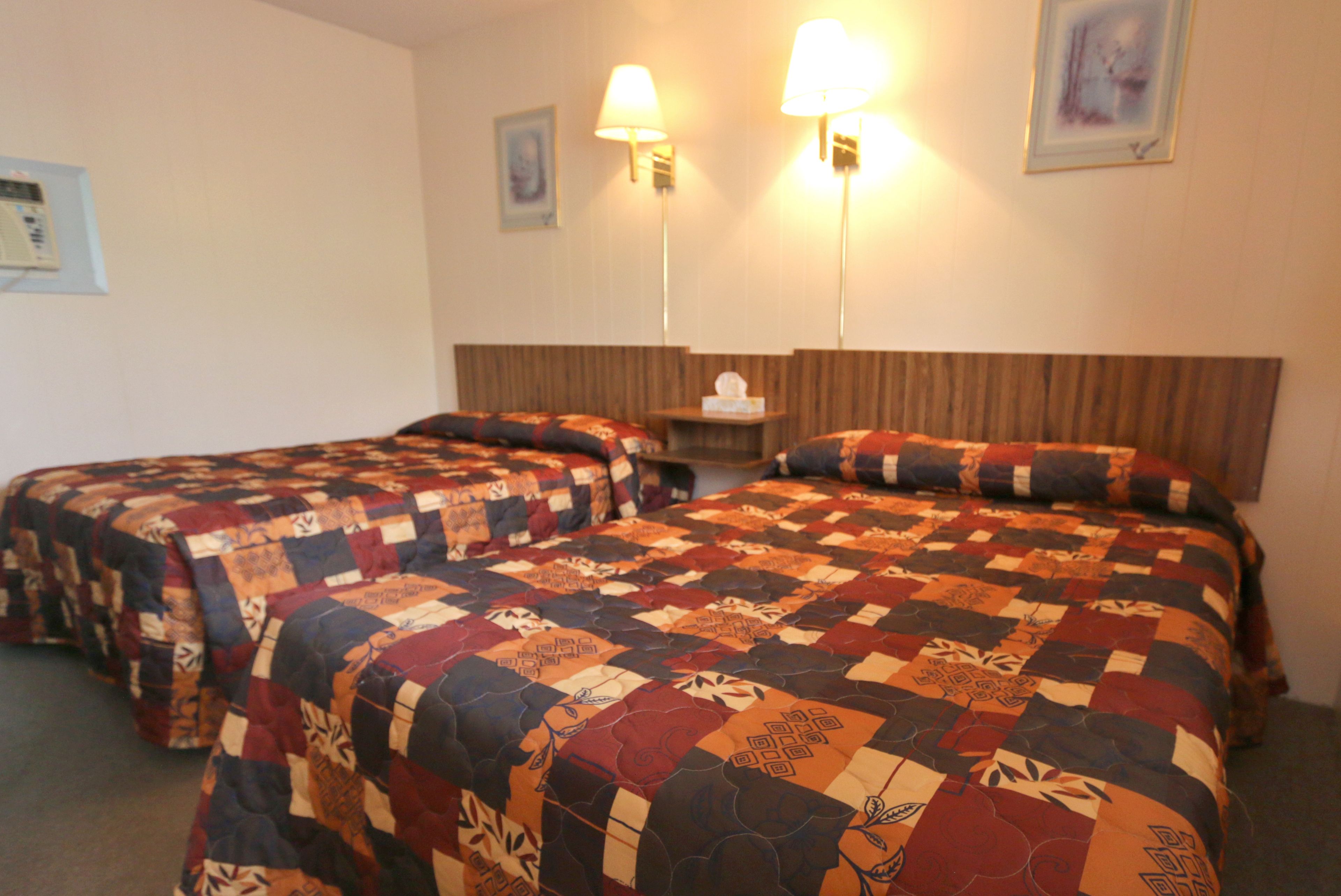 A hotel room with two beds, each covered with a patterned quilt. There are two wall-mounted lamps and a small table with a tissue box.