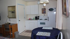 Une cuisine compacte avec des meubles blancs, un four à micro-ondes et une cuisinière. On y trouve une petite table à manger recouverte d’une nappe bleue et agrémentée de sets de table. Une chaise est placée à la table, et une poubelle est visible près de la porte.