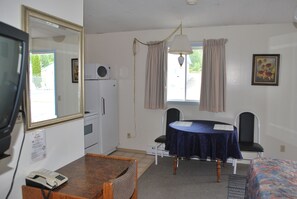 A room with a desk, two chairs, a table with a tablecloth, a bed, a mirror, a window with curtains, and a ceiling light.