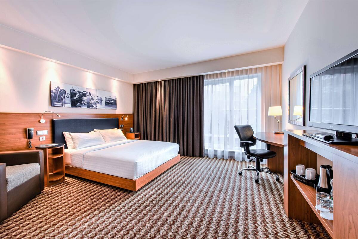 Ett hotellrum med en säng, ett skrivbord, en stol, en tv och ett stort fönster med gardiner.