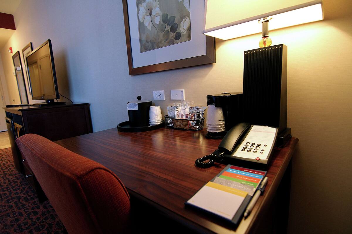 Une chambre d’hôtel avec un bureau en bois, un téléphone, une lampe, un tableau encadré au mur et une chaise rouge.