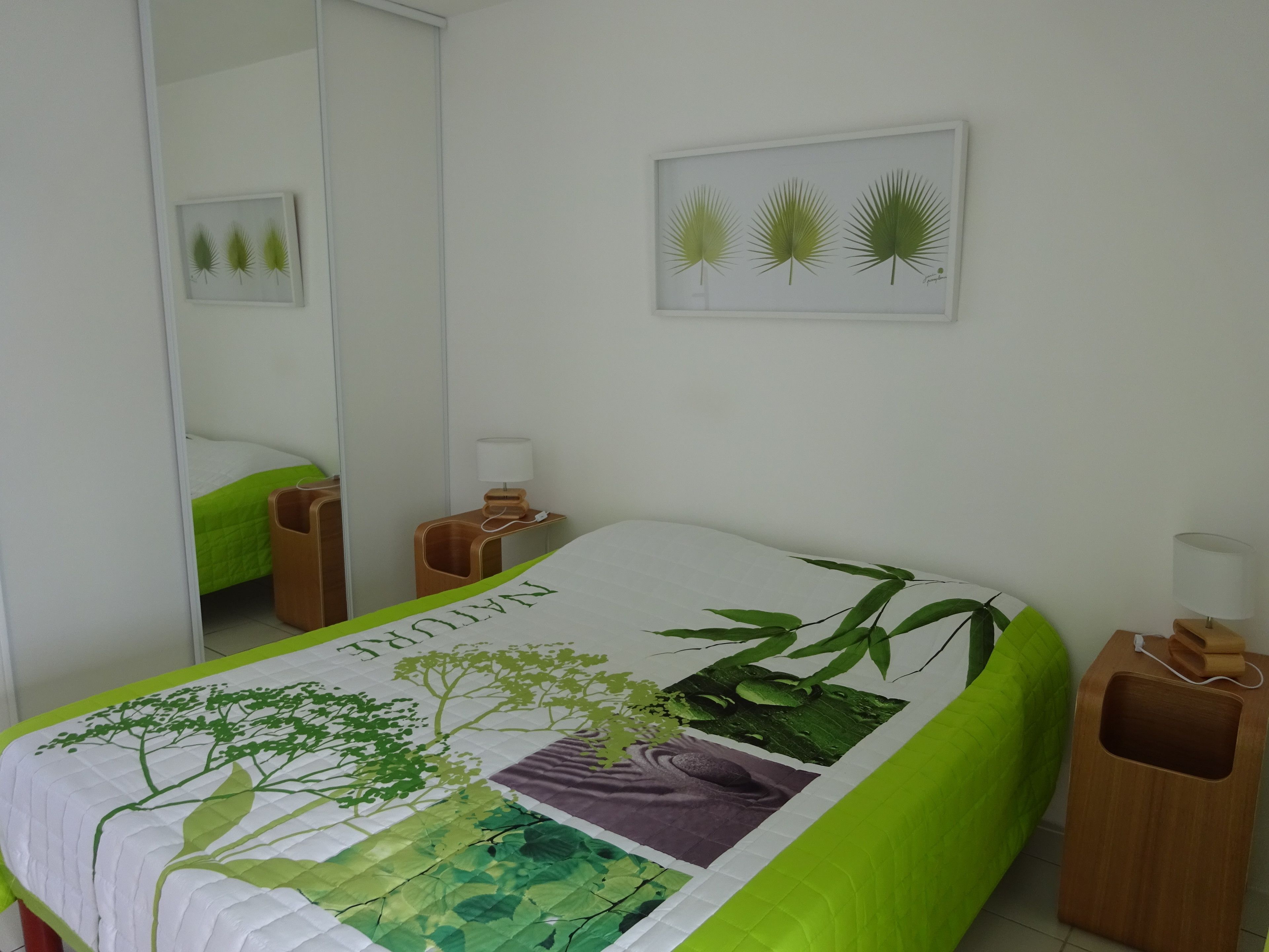 Ein ordentlich bezogenes Bett mit einer grün-weißen Tagesdecke mit Naturmotiv, ein Nachttisch mit Lampe und ein gerahmtes Bild an der Wand.