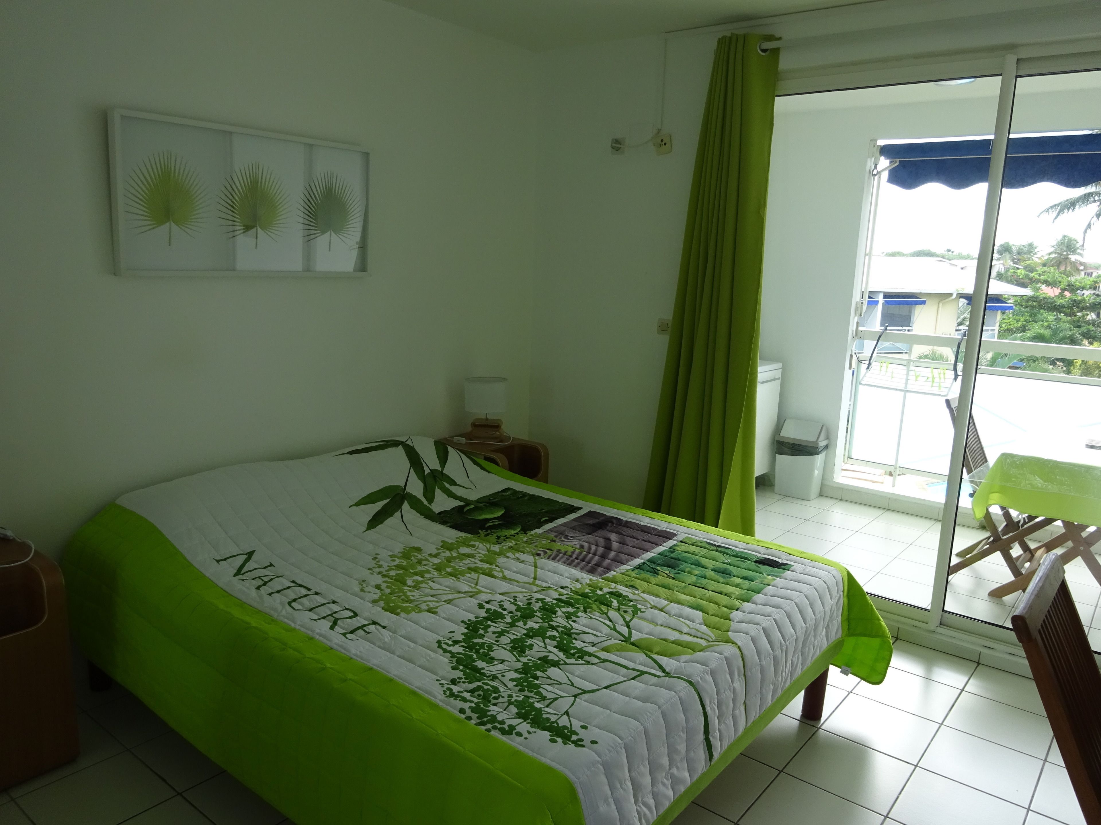 Un dormitorio con cama, colcha con motivos naturales, mesita de noche con lámpara, cortinas verdes y vistas al balcón exterior.