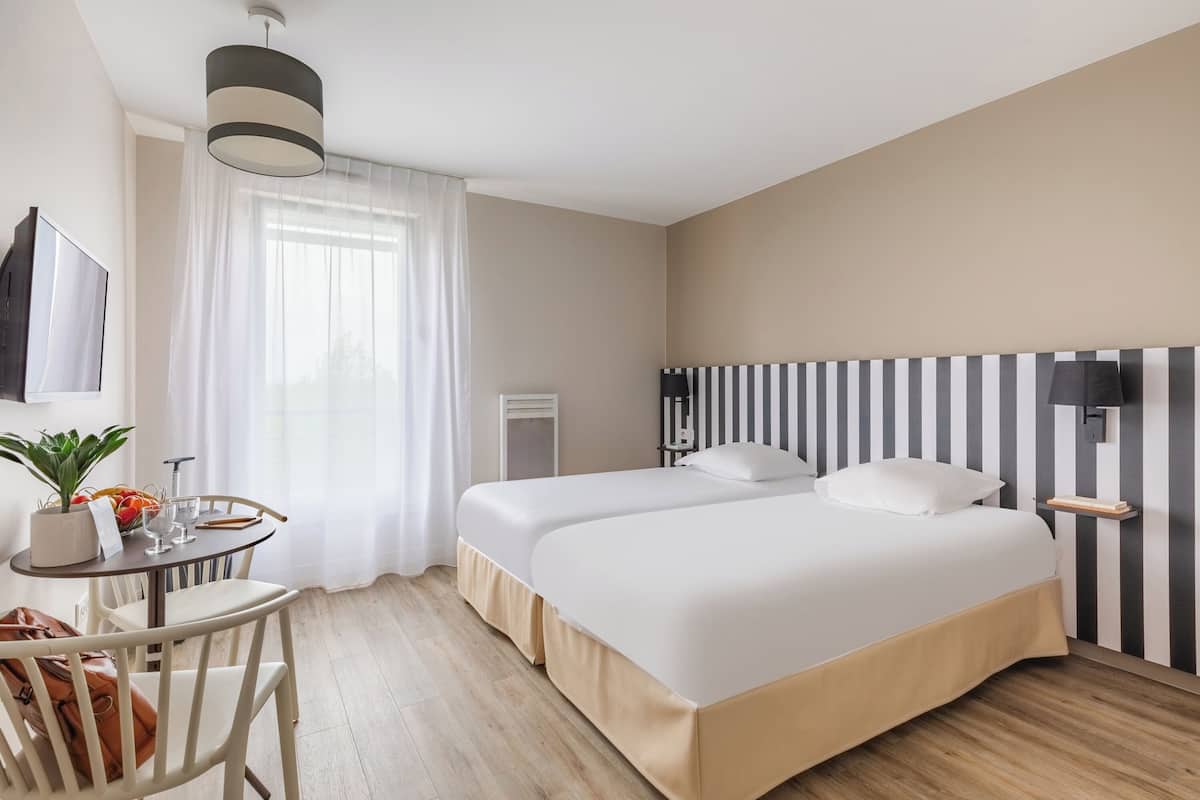 A hotel room with two beds, a desk with a chair, a potted plant, and a TV.
