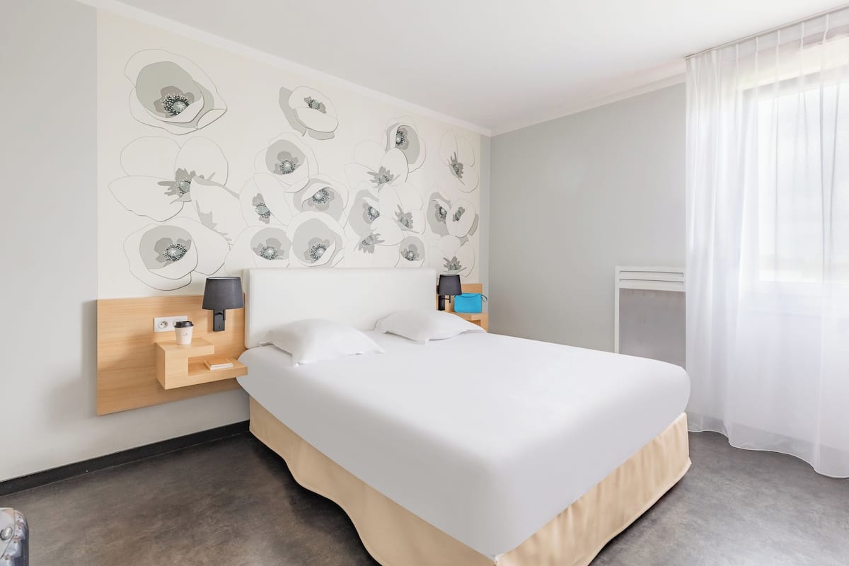 A hotel room with a large bed, a bedside table with a lamp, a wall-mounted artwork, and a window with sheer curtains.