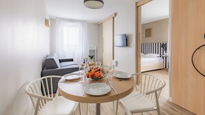 Una habitación moderna y bien iluminada con una mesa redonda de comedor preparada para dos personas, un sofá, un televisor y una cama con ropa de cama a rayas.