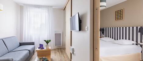 Una habitación de hotel moderna con un sofá gris, una pequeña mesita de madera con una planta, un televisor de pantalla plana montado en la pared y una cama con ropa de cama a rayas.