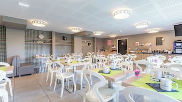 A modern dining area with white chairs and tables, a kitchen in the background, and a ceiling with a unique pattern.
