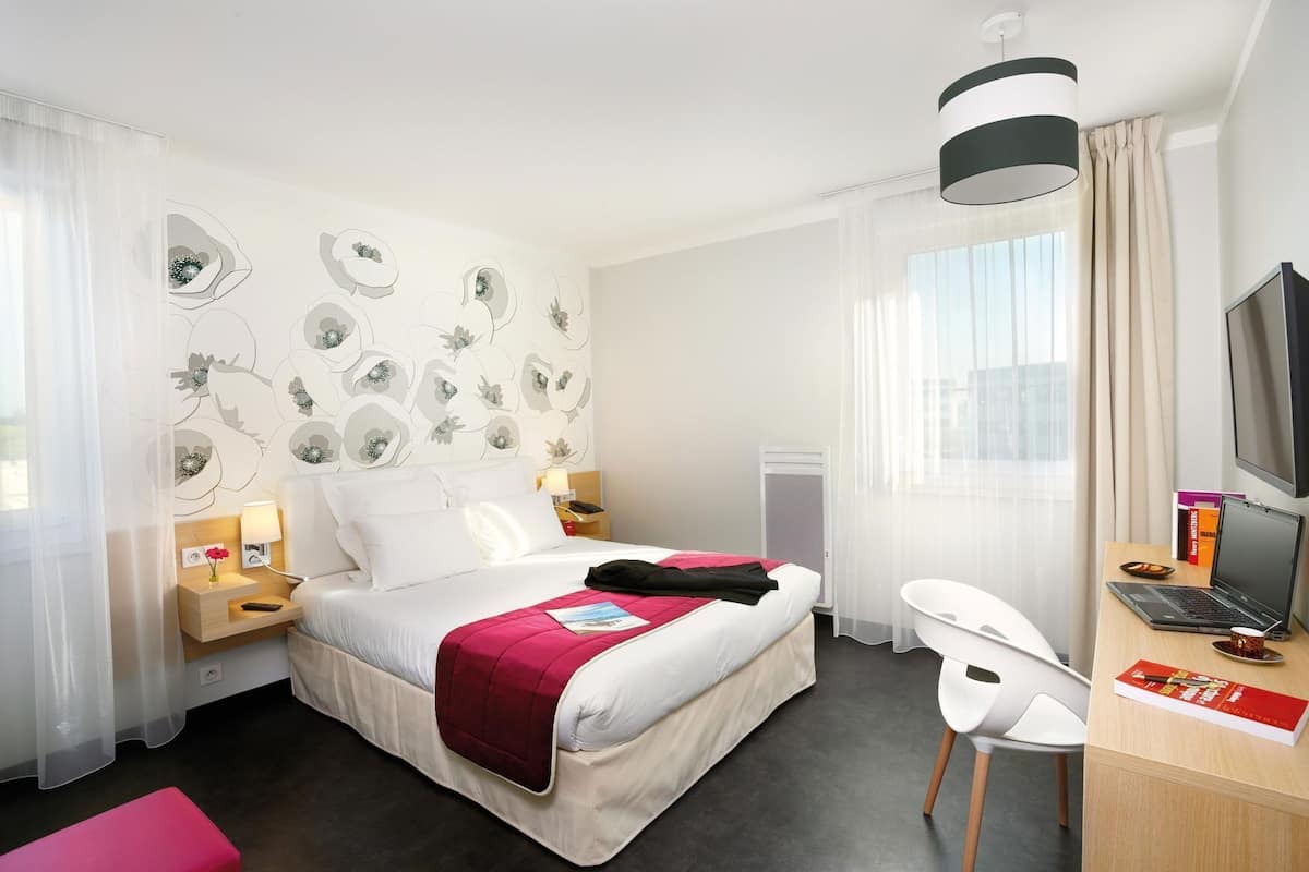 A hotel room with a bed, desk, chair, TV, and a window with curtains.