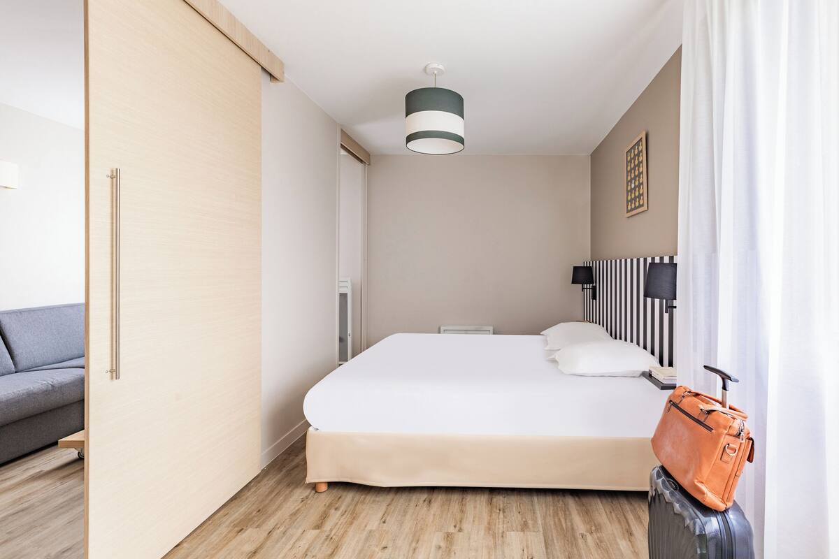 A hotel room with a bed, a grey sofa, a suitcase, and a handbag.