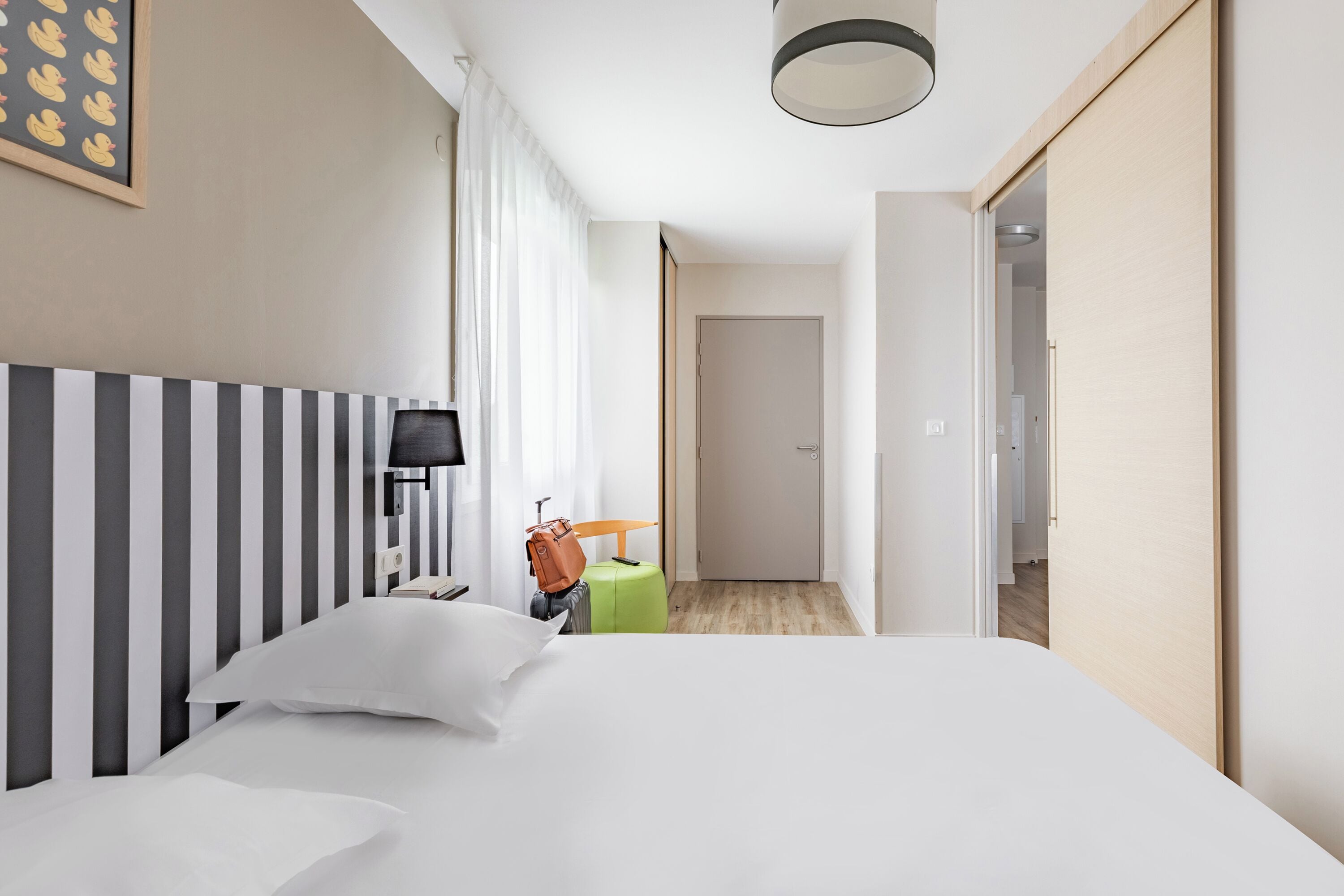 Quarto de hotel com uma cama grande, uma mesa de cabeceira, uma luminária, uma mala e uma porta que dá para outro cômodo.