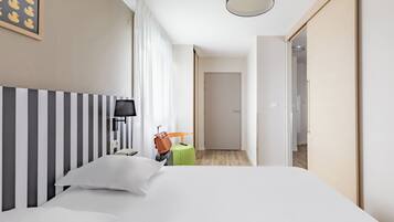 Quarto de hotel com uma cama grande, uma mesa de cabeceira, uma luminária, uma mala e uma porta que dá para outro cômodo.