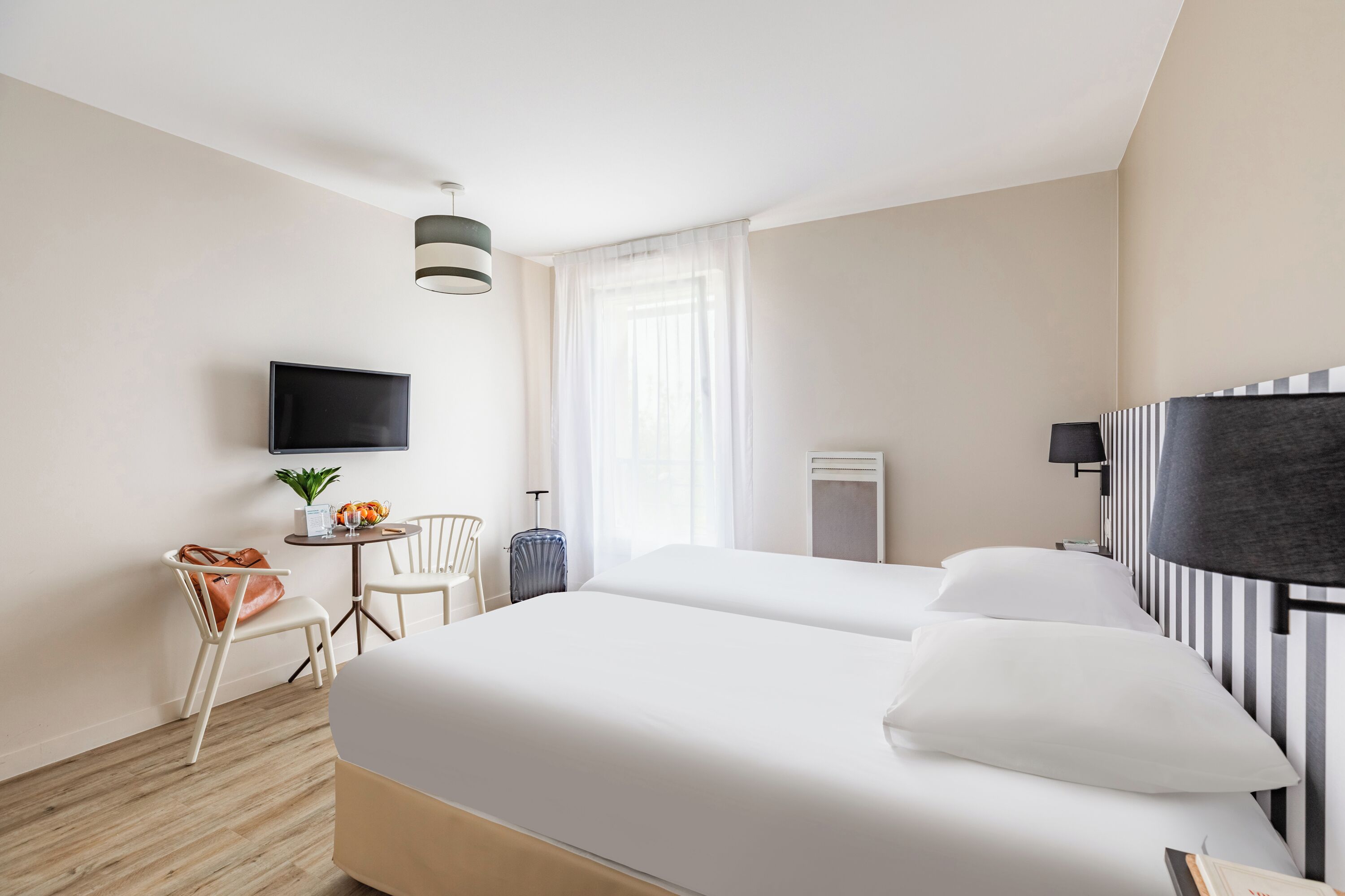 Habitación de hotel con dos camas, un televisor montado en la pared, una mesita con una planta, una silla y una maleta.