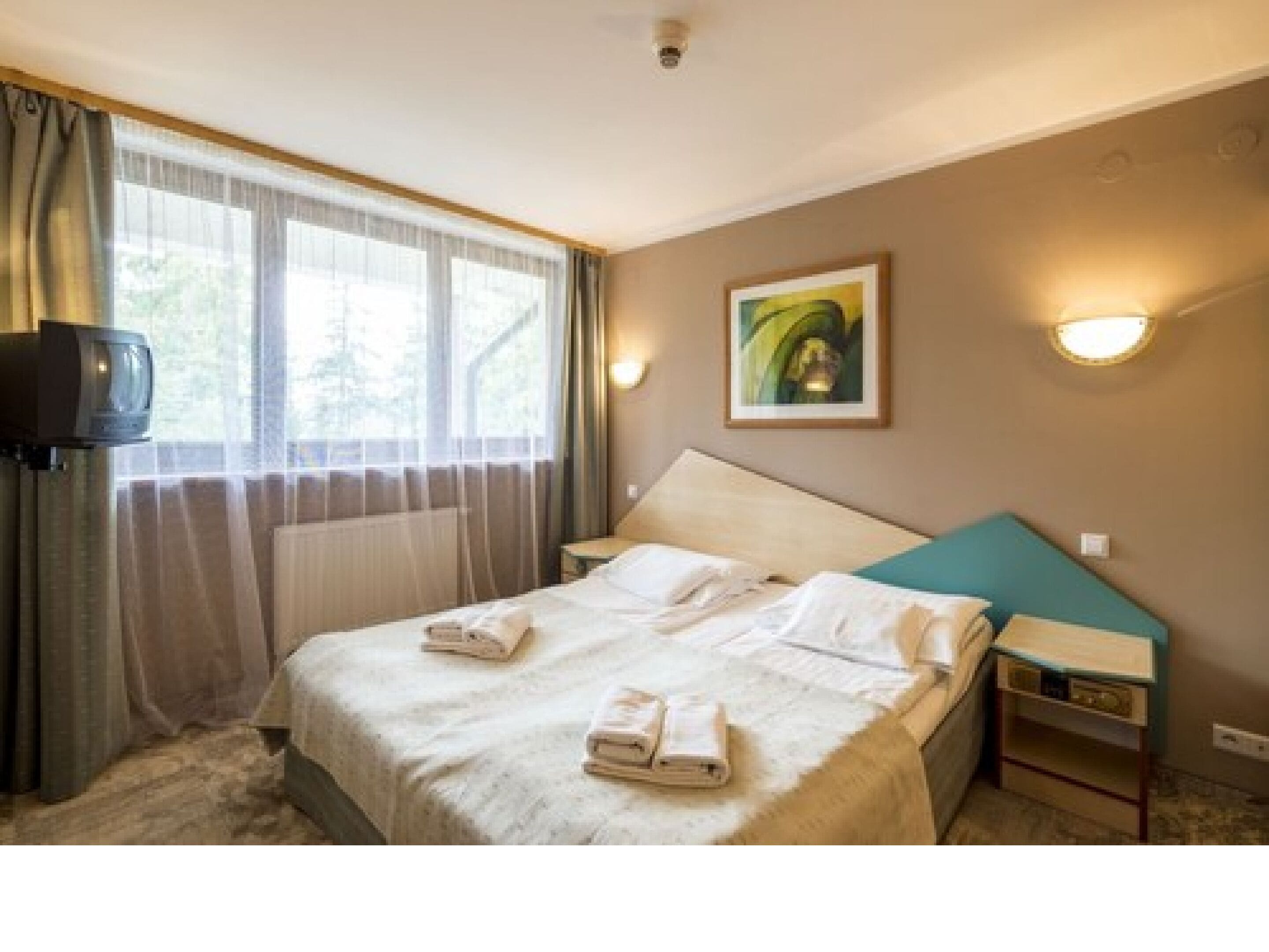 A hotel room with a bed, a bedside table, a TV, and a painting on the wall.