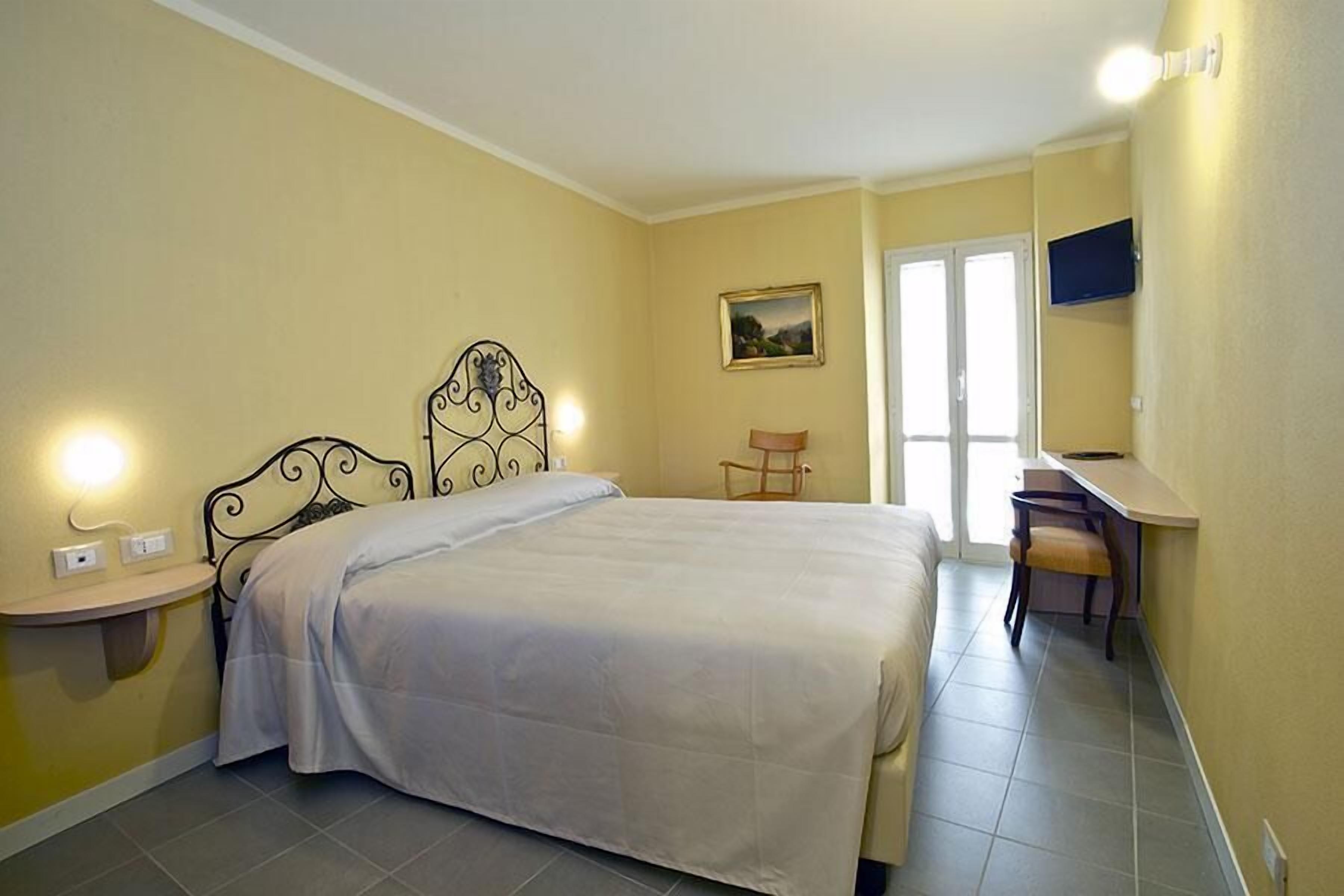 Un dormitorio con cama, mesita de noche, silla, escritorio y un cuadro en la pared.