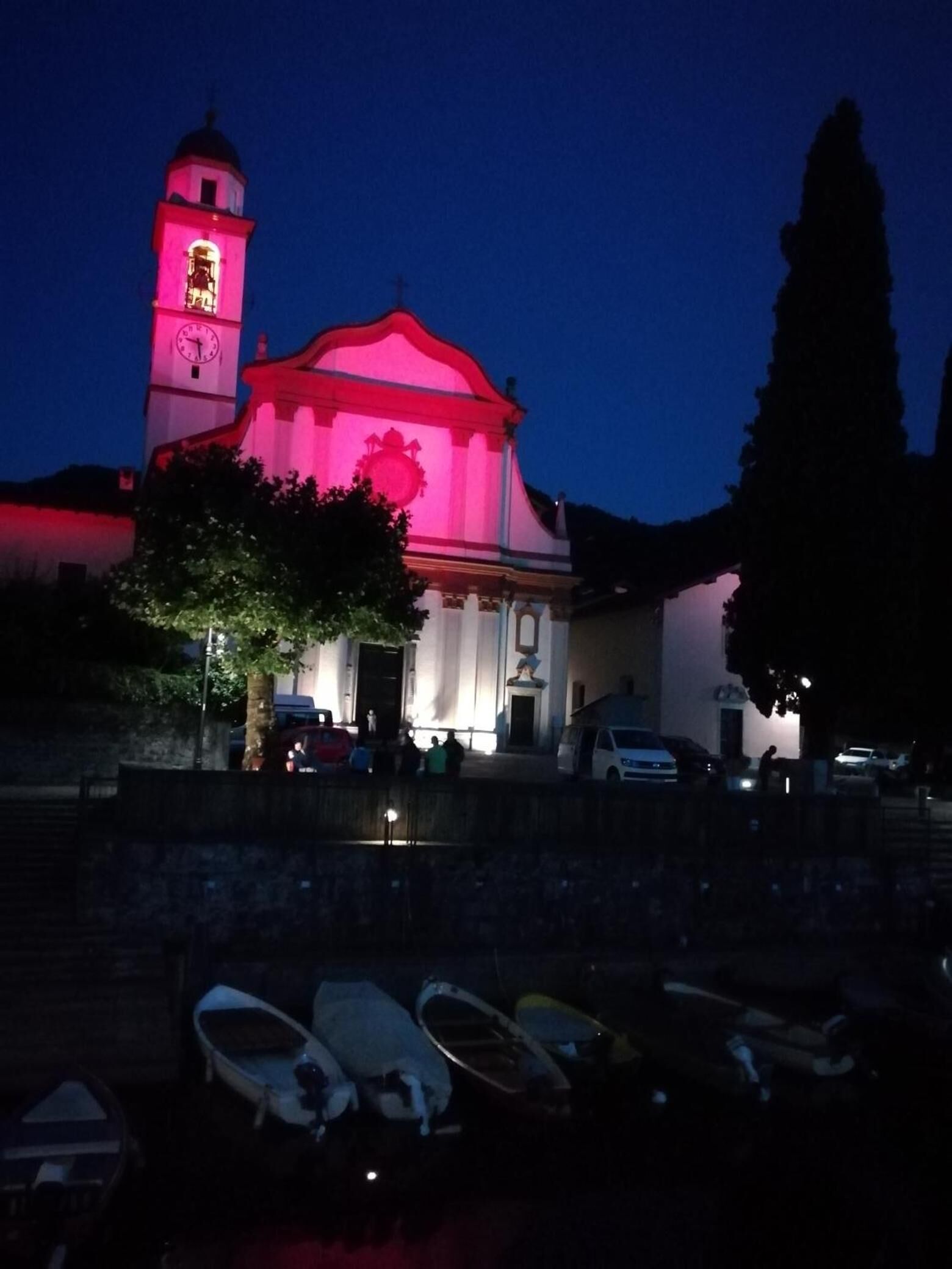 Uma igreja com uma torre de relógio, telhado vermelho e um sino. Há barcos em primeiro plano.
