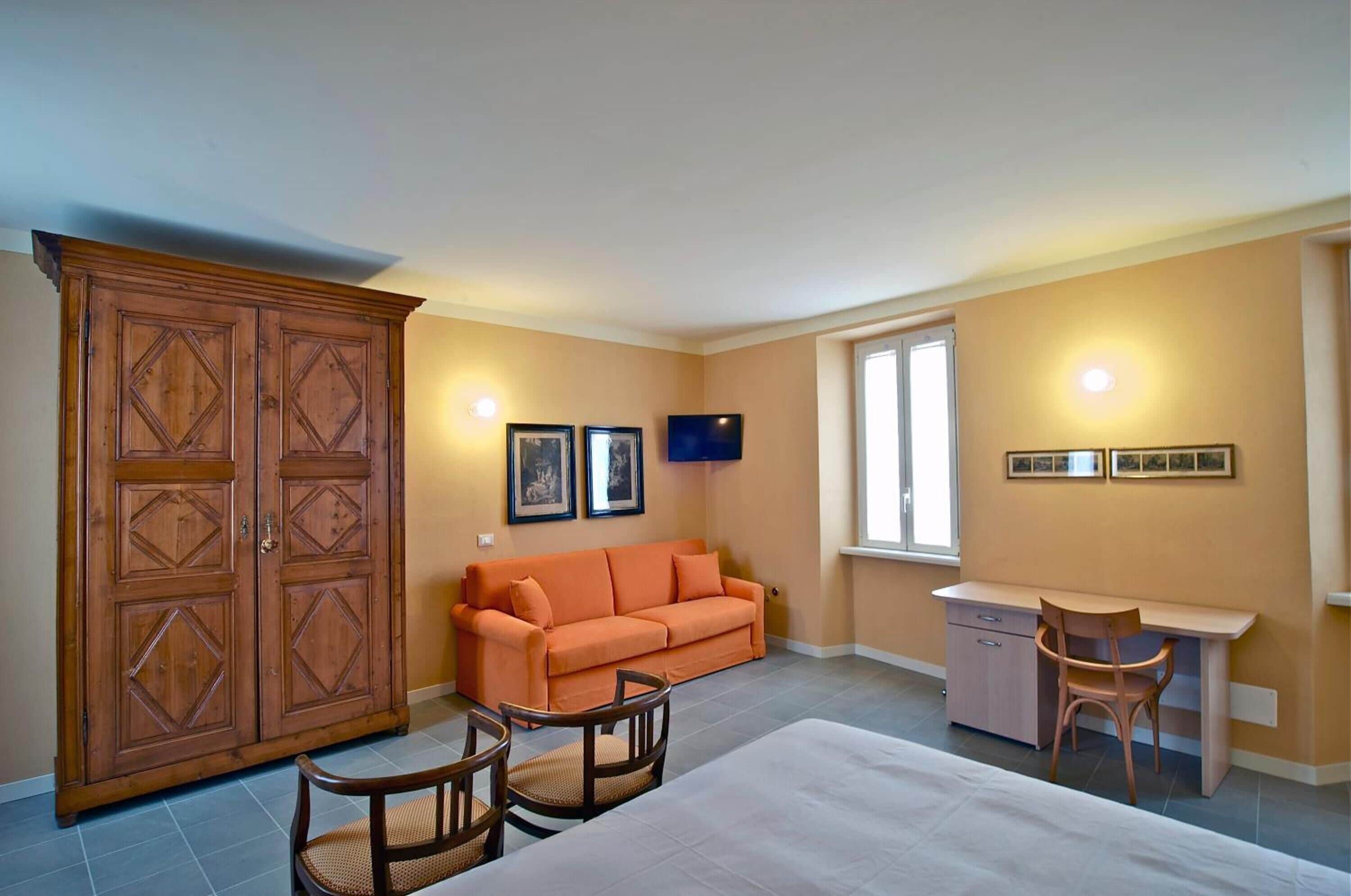 Una habitación con un armario grande de madera, un sofá naranja, una pequeña mesa de comedor con dos sillas, un escritorio con una silla y un televisor montado en la pared.