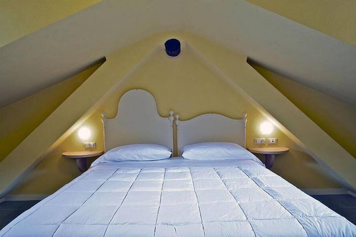 A triangular room with a bed centered under a sloped ceiling, two bedside lamps, and a small round window at the peak.