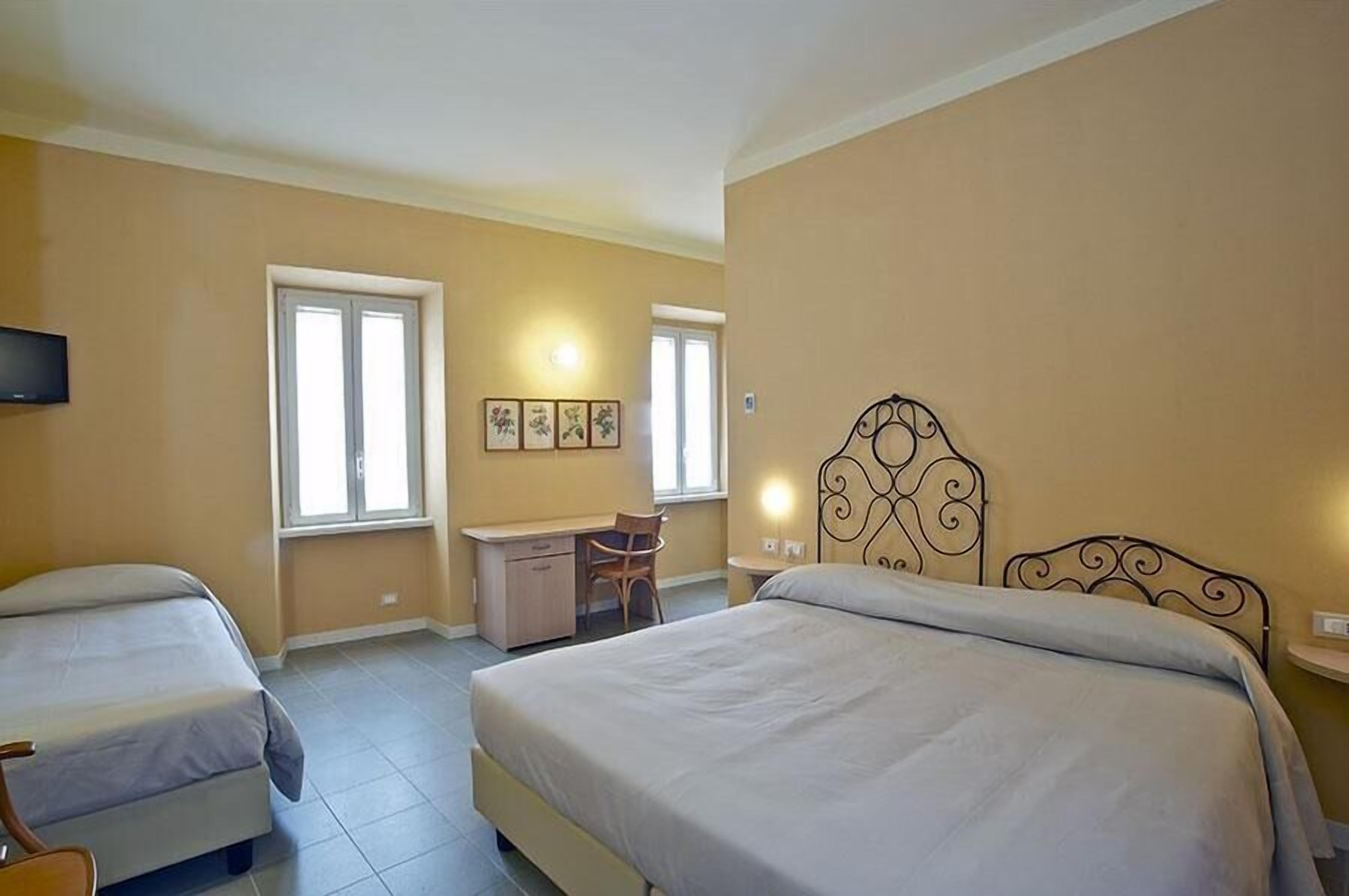 A hotel room with two beds, a desk, and a chair. There are two windows and wall-mounted artwork.
