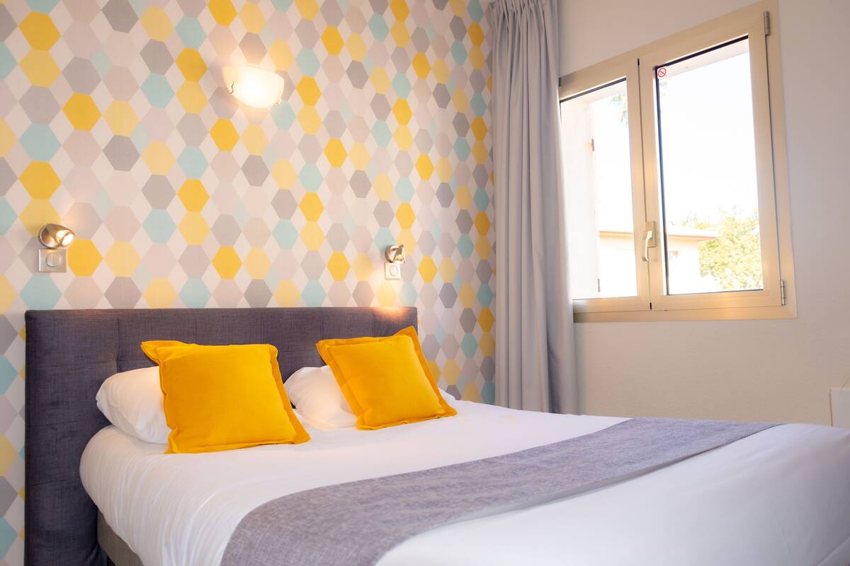 Un lit bien fait, avec une tête de lit grise et deux coussins jaune vif, placé devant un mur recouvert d’un papier peint à motifs géométriques.