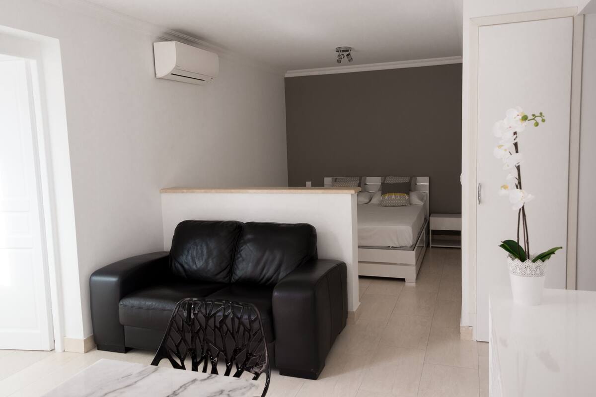 Un espace de vie moderne et compact, comprenant un canapé en cuir noir, une petite table à manger et une plante orchidée blanche.