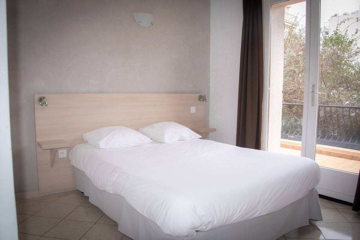 Un lit simple avec une tête de lit en bois, une literie blanche et une vue sur l’extérieur grâce à une grande fenêtre.