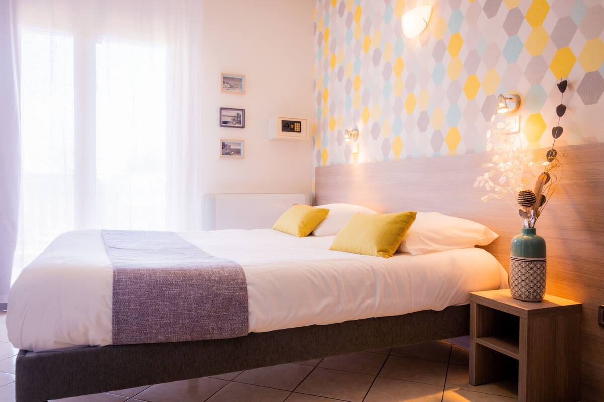 Un lit bien fait, orné d’oreillers blancs et jaunes, une table de chevet avec un vase décoratif, et un mur recouvert d’un papier peint à motifs géométriques.