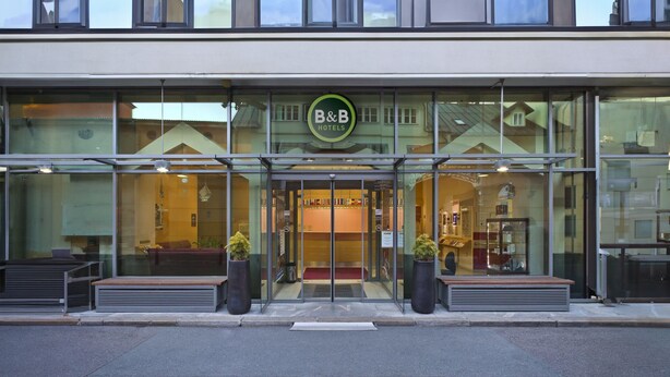 A B&B hotel entrance with a glass facade, a green sign, and two potted plants on the sidewalk. - B&B HOTEL Maribor UNI Hostel (Maribor)