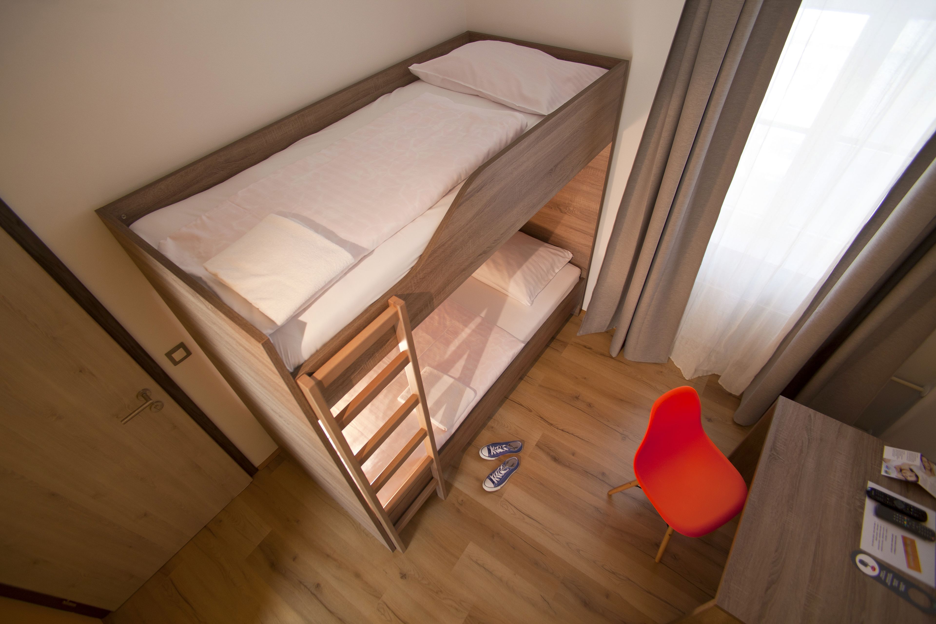 Uma cama beliche com escrivaninha e cadeira, piso de madeira e janela com cortinas.