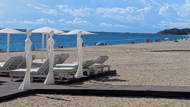 Una playa con sillas de sol blancas y sombrillas, un paseo marítimo de madera y el mar al fondo.