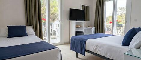 Habitación de hotel con dos camas, televisión y balcón con vistas a árboles y flores.
