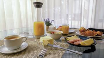 Ein Frühstückstisch mit Orangensaftgläsern, Obstschale, Croissants, Käse und Schinken auf einer Glastischplatte.
