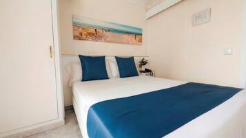Una cama bien hecha con una colcha azul y almohadas, un cuadro de una playa en la pared, y una mesita de noche con un teléfono.