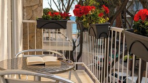 Un balcón con una silla de metal, una mesa con un libro abierto y flores rojas en macetas.