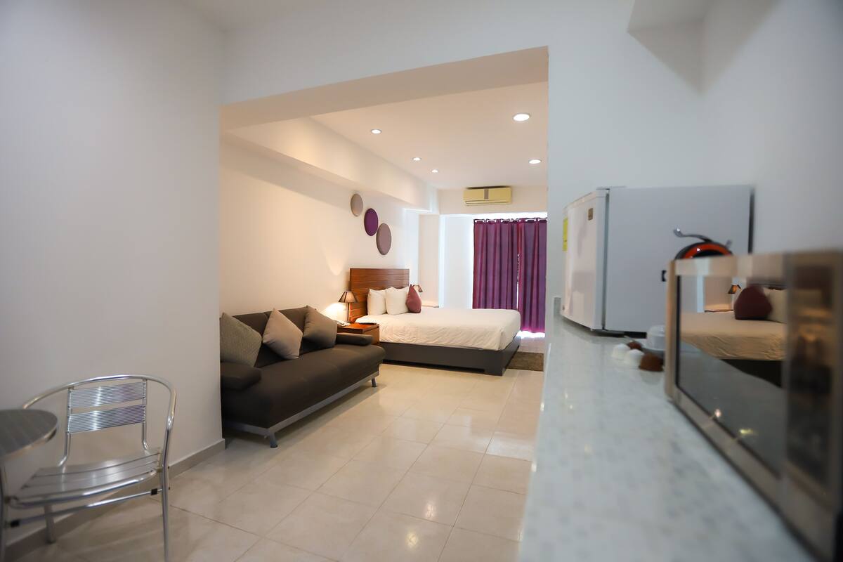 A modern hotel room with a bed, sofa, and a dining area.
