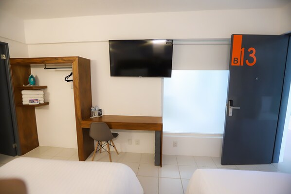 Quarto de hotel com cama, escrivaninha, cadeira, televisão e guarda-roupa.