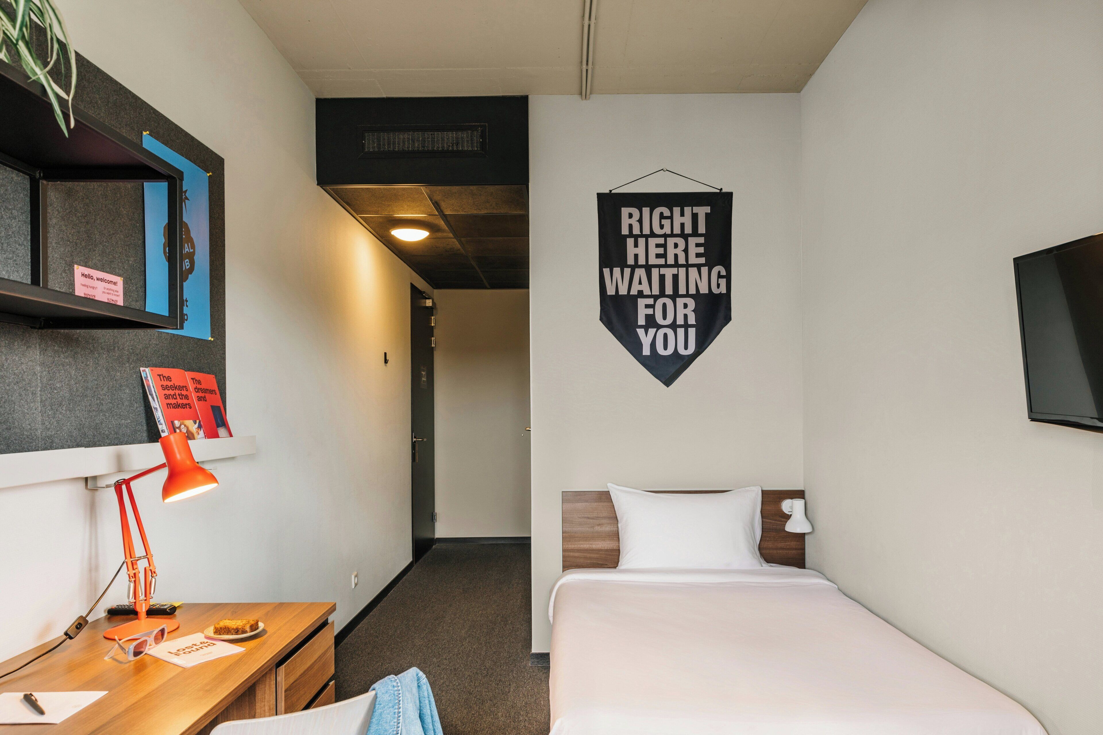 Habitació d'hotel amb un llit, una taula de nit amb una làmpada, un televisor penjat a la paret i un cartell que diu "RIGHT HERE WAITING FOR YOU".