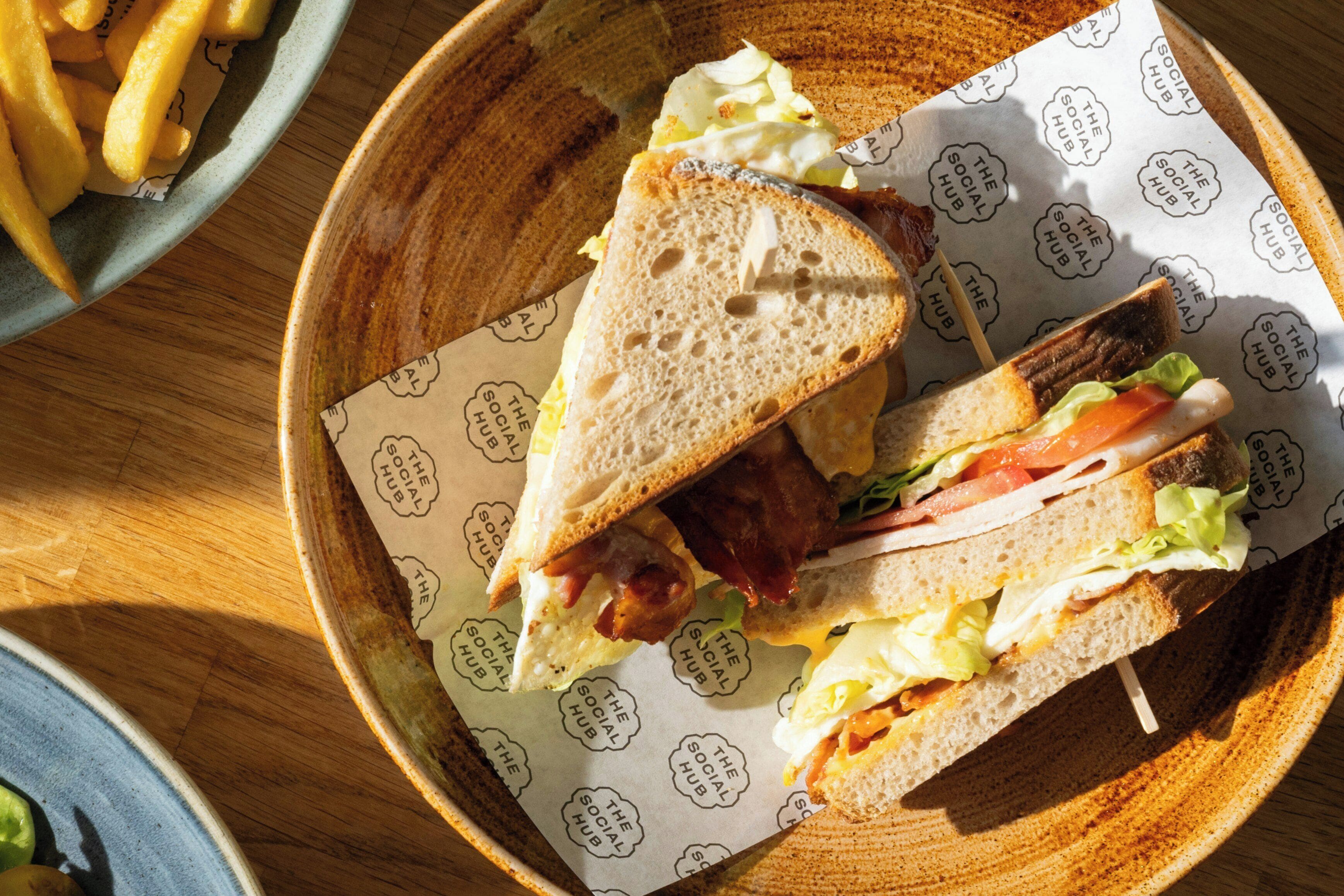 Un panino con lattuga, pomodoro e bacon su una fetta di pane, appoggiato su carta con un logo.