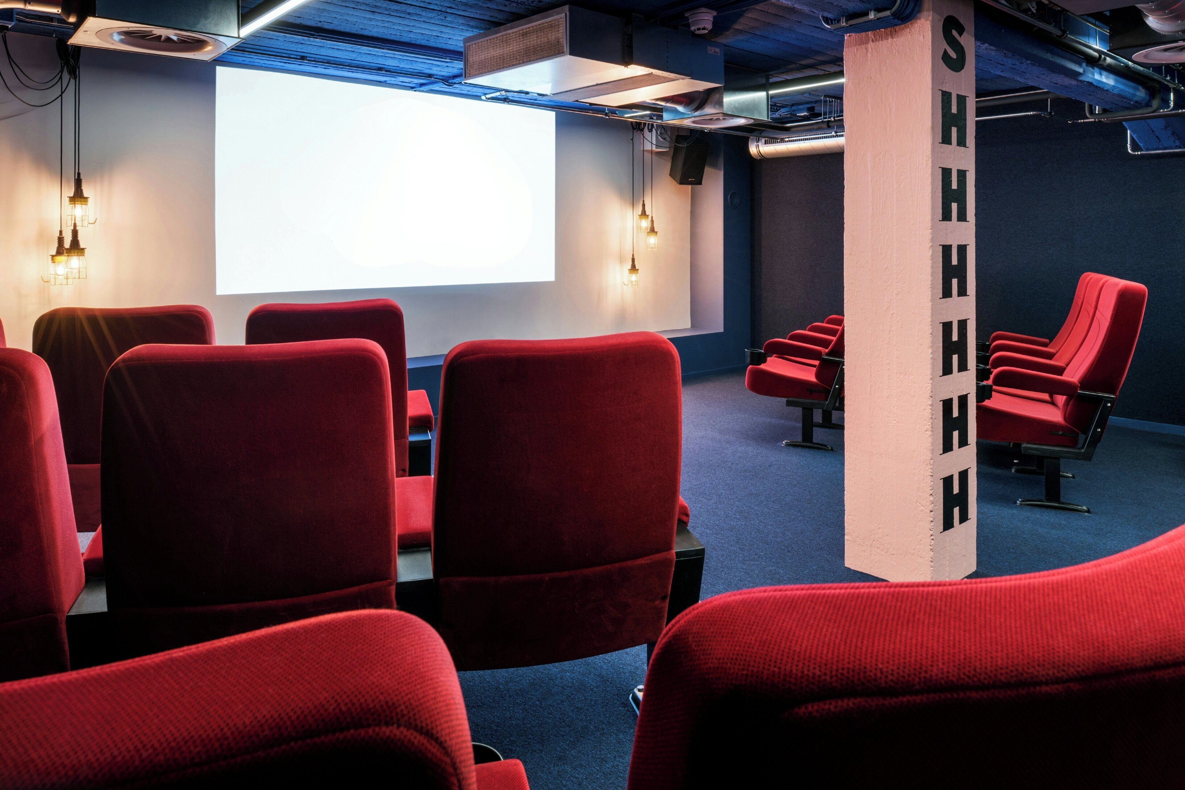 Una sala de cine con asientos rojos, una pantalla grande y un pilar con la leyenda 'SHHHH'.