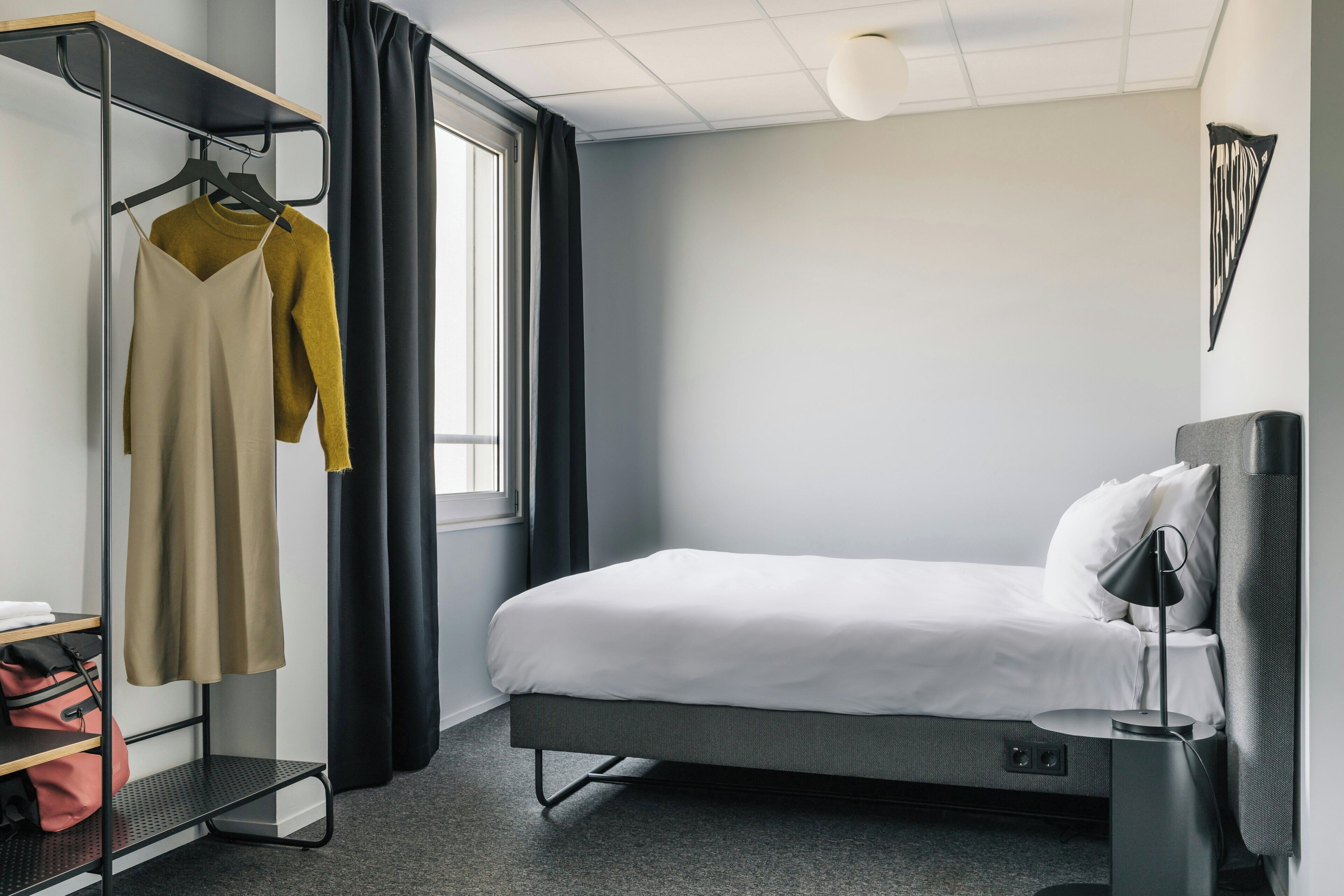 A hotel room with a bed, a bedside table with a lamp, a wardrobe, and a window with curtains.