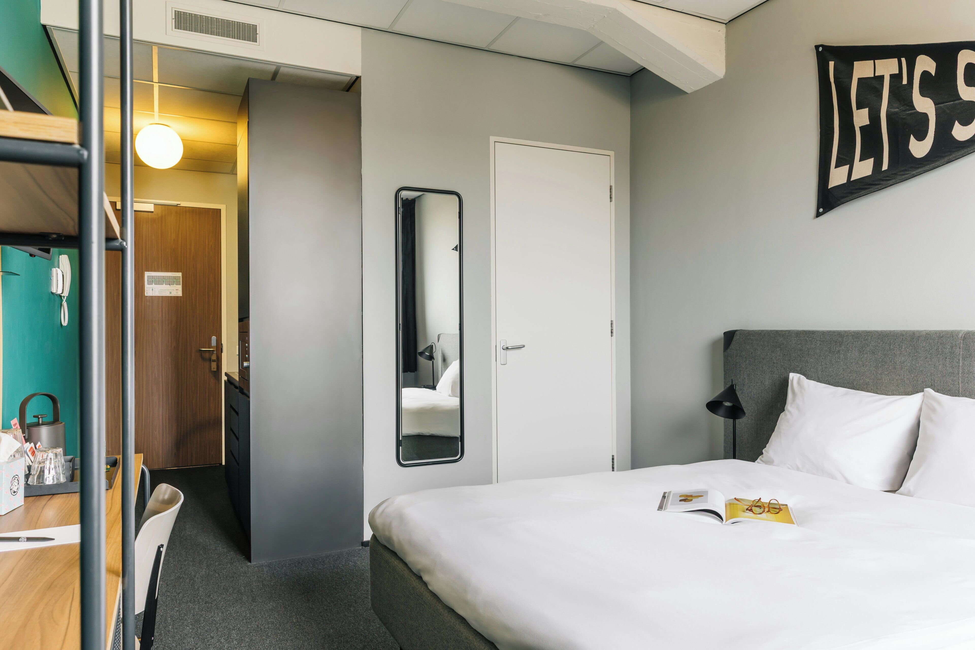 A hotel room with a bed, a bedside table, a mirror, a door, and a sign on the wall.