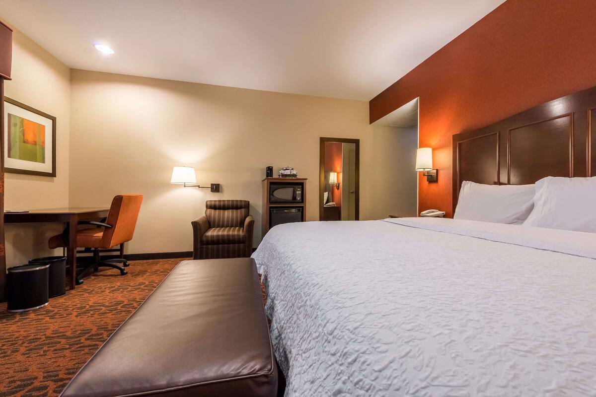 A hotel room with a large bed, a desk with a chair, a small armchair, a trash bin, and a mirror.