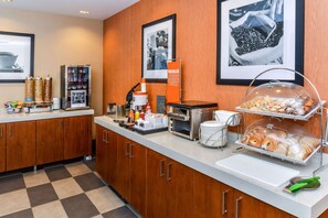 A hotel breakfast area with a coffee machine, condiment dispensers, a coffee urn, a juice dispenser, and a pastry display.