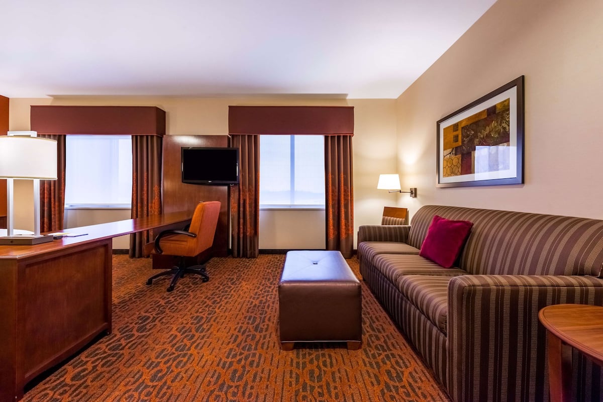 A hotel room with a desk, chair, sofa, ottoman, and a TV mounted on the wall.