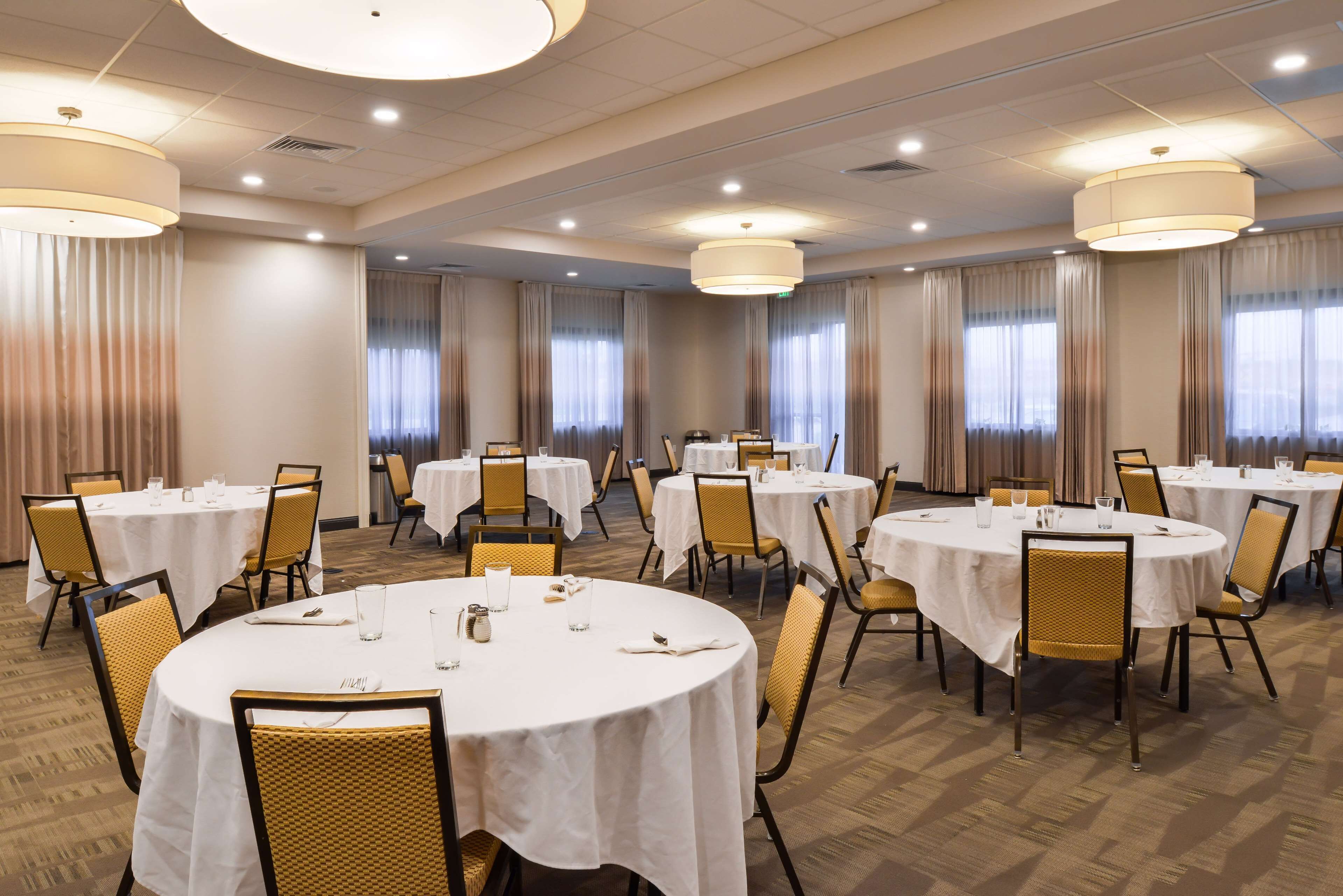 Un salón de conferencias de hotel con varias mesas redondas, sillas y ventanas amplias con cortinas.