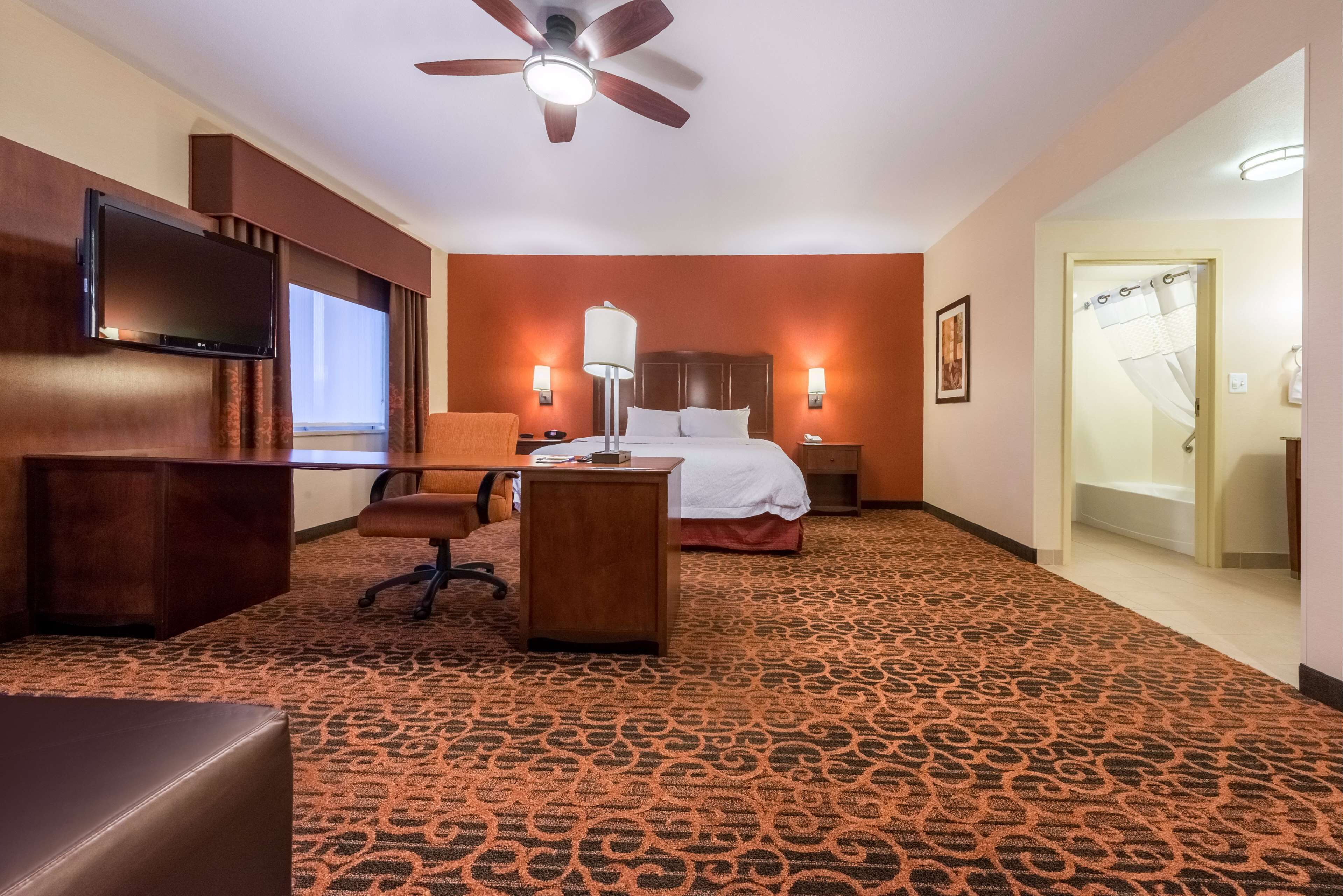 Habitación de hotel con cama, escritorio, silla, televisión y ventilador de techo.