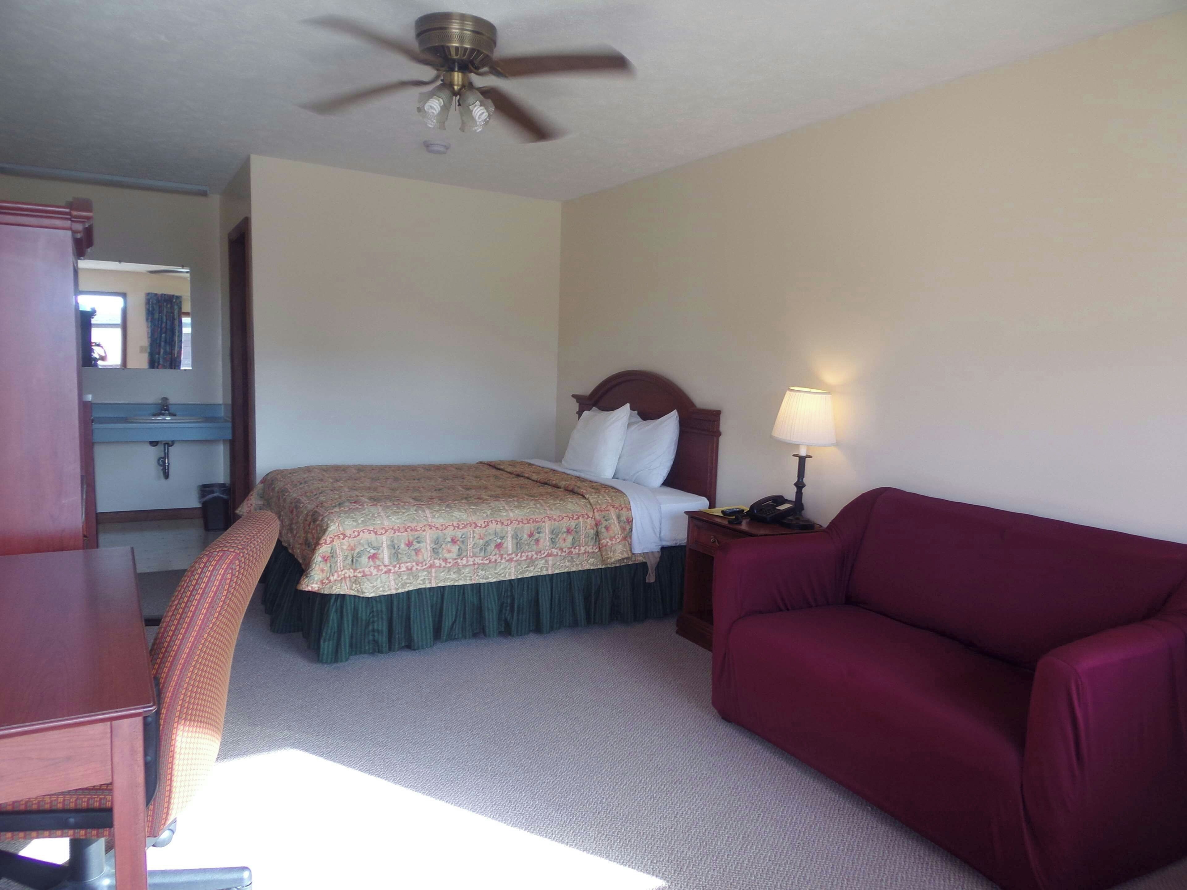 Habitación de hotel con cama, escritorio, silla, sofá, mesita de noche con lámpara, ventilador de techo y baño visible a través de una puerta abierta.